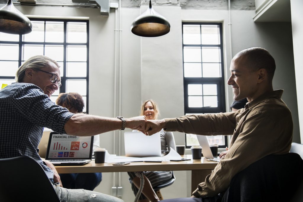 Two men smiling and fist bumping in an office with people in the background for Wellspace blog Looking After Your Workplace Wellbeing